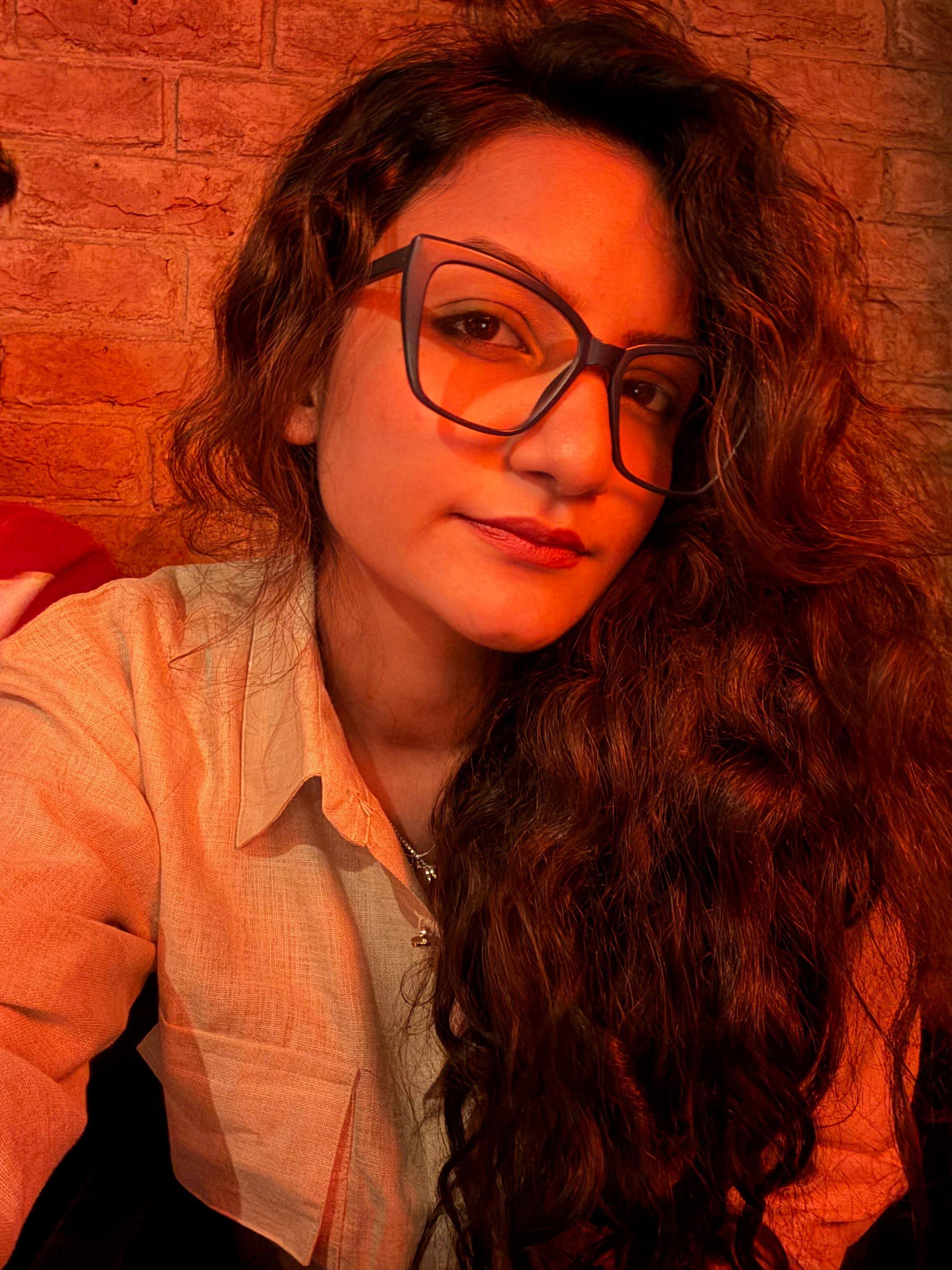 Preet Kaur with a slight smile, wearing large glasses, with long curly hair, in warm orange light against a brick wall